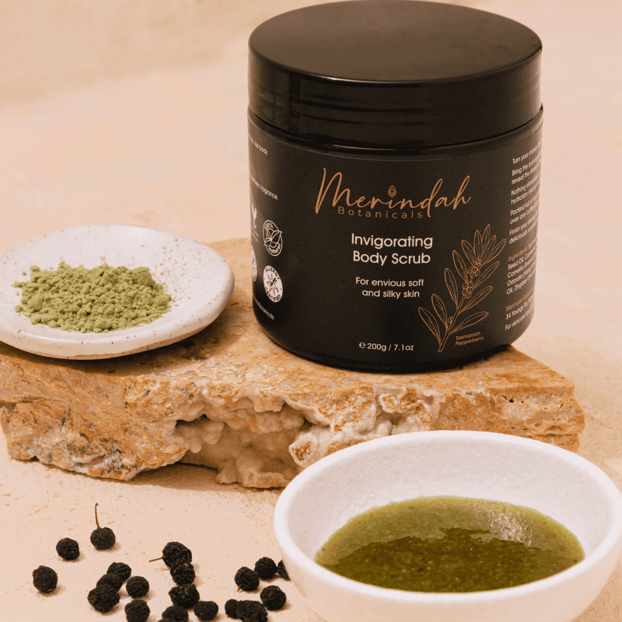 A small jar of Merindah Botanicals Invigorating Body Scrub is displayed on a stone surface. Nearby are two white bowls, one containing all-natural green powder and the other a green paste, with black peppercorns scattered around.