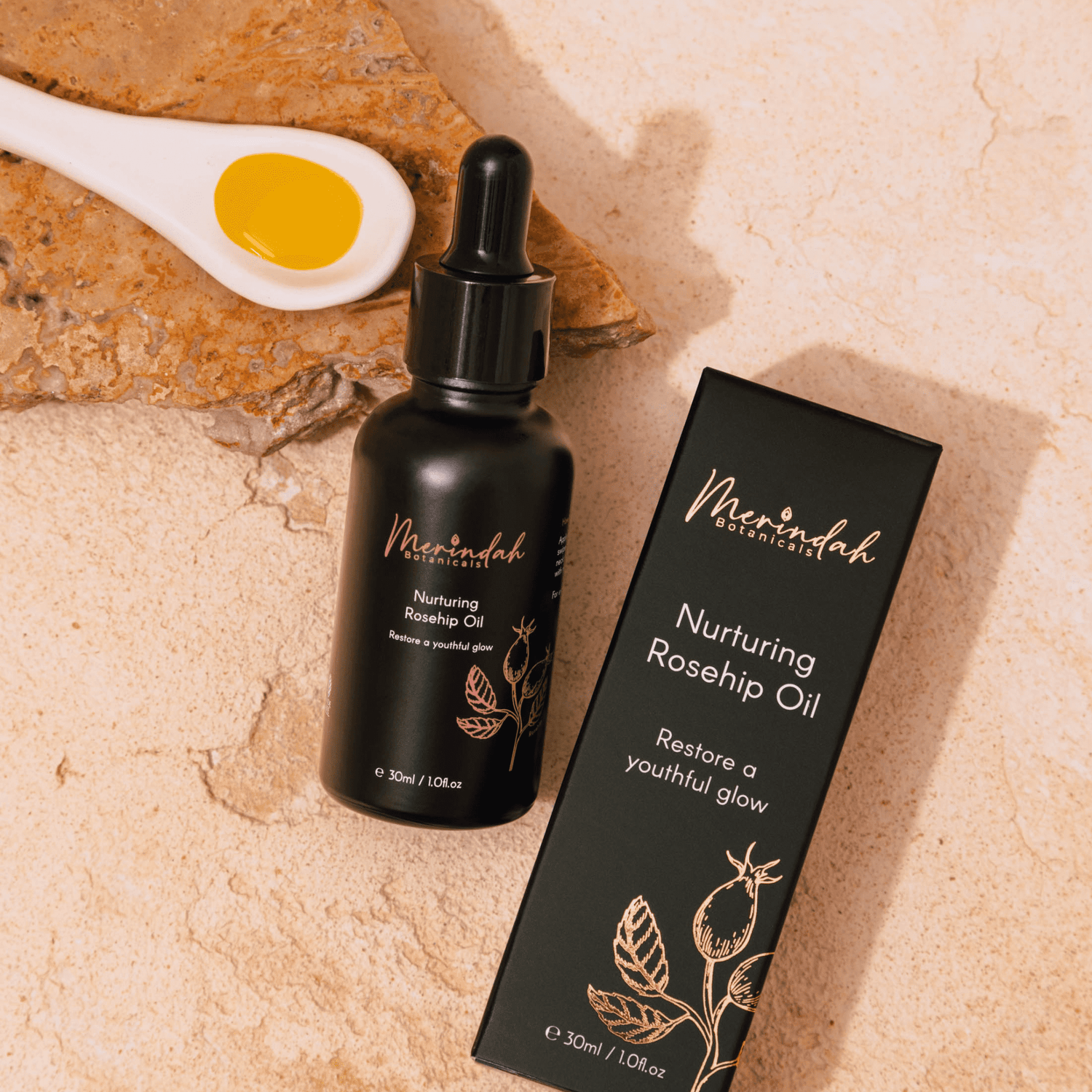 A bottle of Merindah Botanicals certified organic Rosehip Oil with its packaging box, displayed on a stone surface on a small ceramic spoon 
filled with oil beside it.