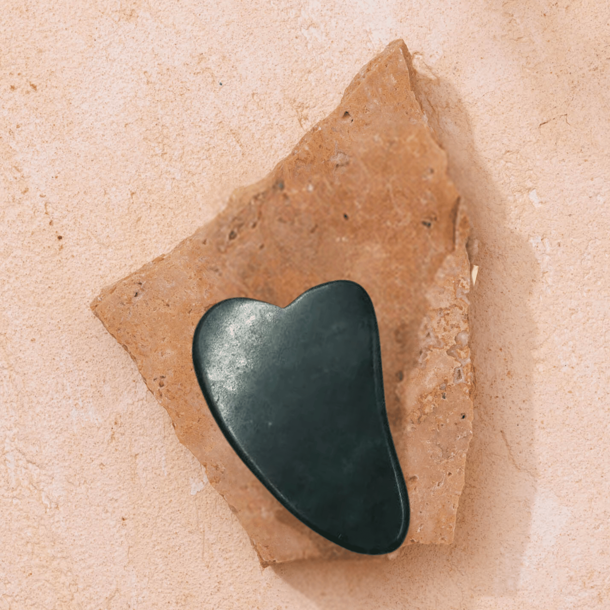 A heart-shaped Gua Sha tool from Merindah Botanicals sits elegantly on a rugged, irregular piece of beige stone against a complementary textured background, offering a revitalizing facial experience as an integral part of your skincare regimen.