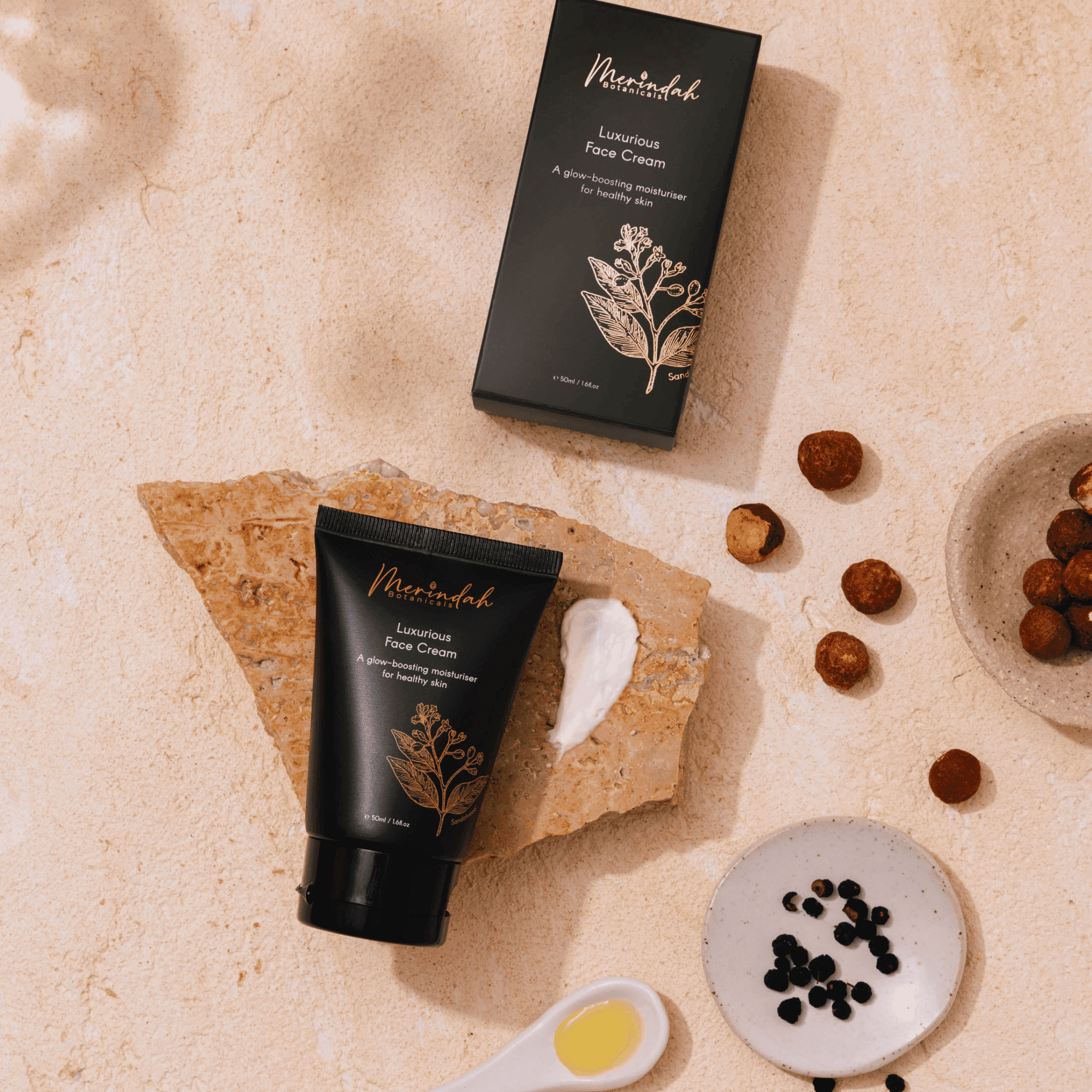 Flatlay image of Merindah Botanicals Luxurious Face Cream on beige background with natural skincare ingredients arranged next to the black box and tube.