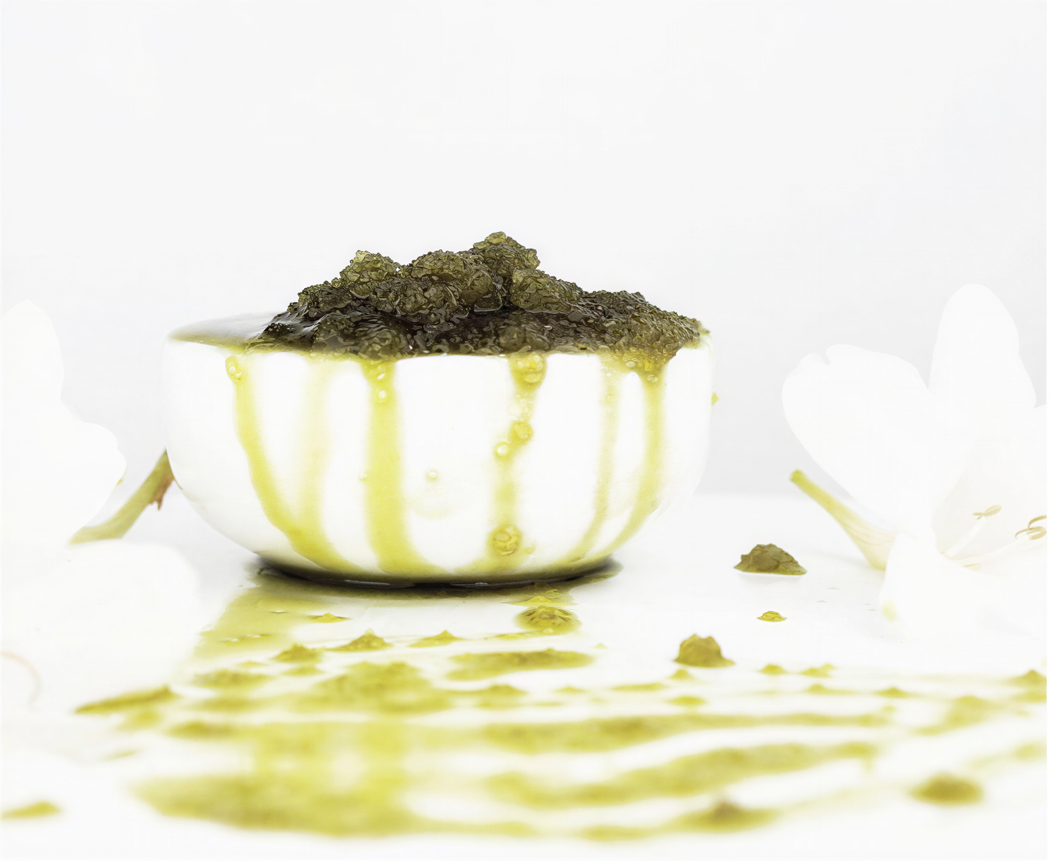 A white bowl brimming with Invigorating Body Scrub by Merindah Botanicals rests on a surface. The all-natural green scrub cascades over the edges, dripping onto the pristine white background, surrounded by soft-focus white flowers.