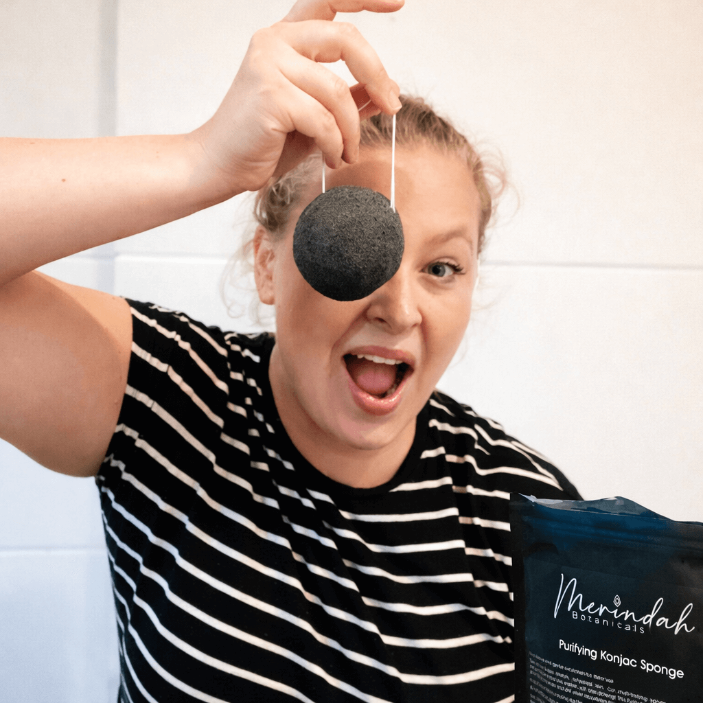 A smiling woman holding a black konjac sponge in front of her face
