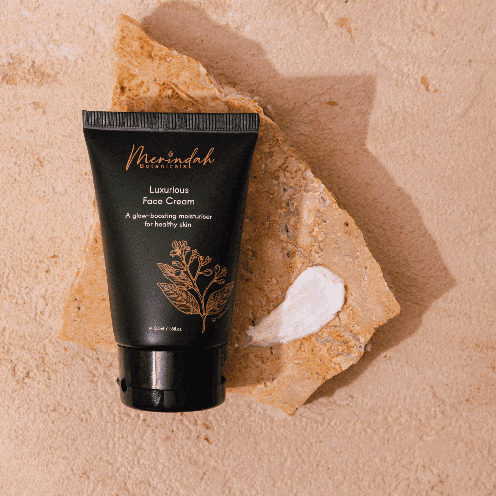 Black tube of Merindah Botanicals face cream arranged on natural sand stone and beige background with a smear of the product to one side.